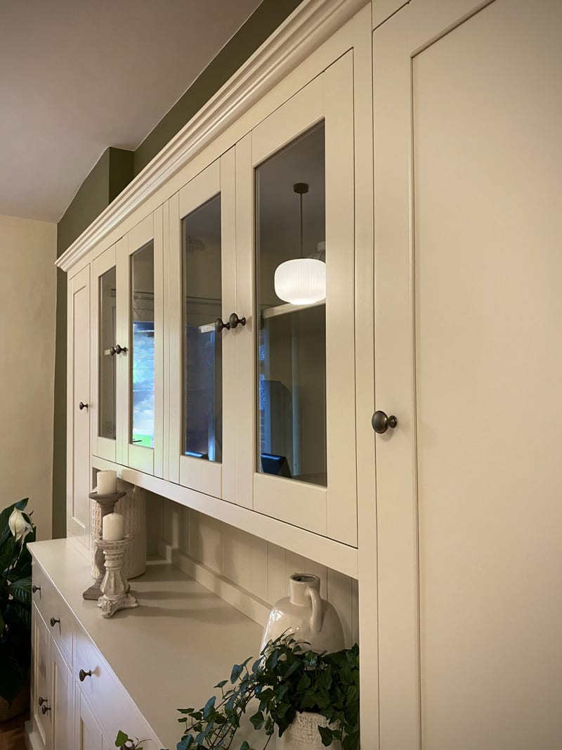 View of the upper section from the right, the glazed doors reflecting a ribbed pendant light, with the tall solid end door, moulded cornice and candlesticks on the worktop below