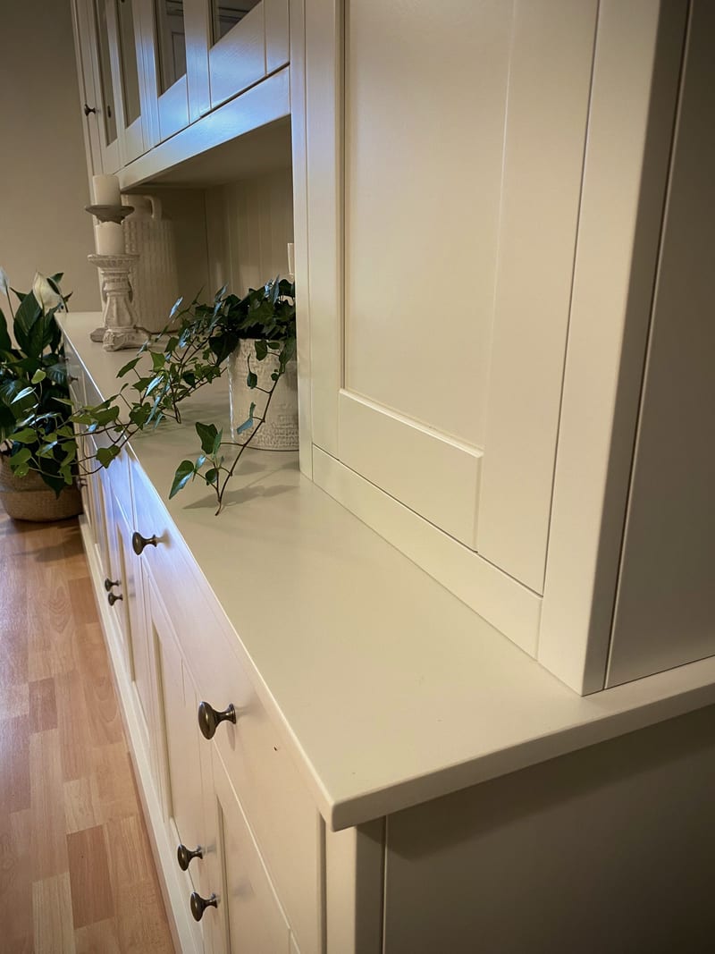 Close view across the painted cream worktop to the solid right-hand door, showing the shaker panel, trailing ivy and the aged bronze knobs on the drawers beneath