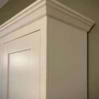 Detail of the top corner, showing the layered moulded cornice and the painted side panel meeting the shaker door against the olive green wall behind