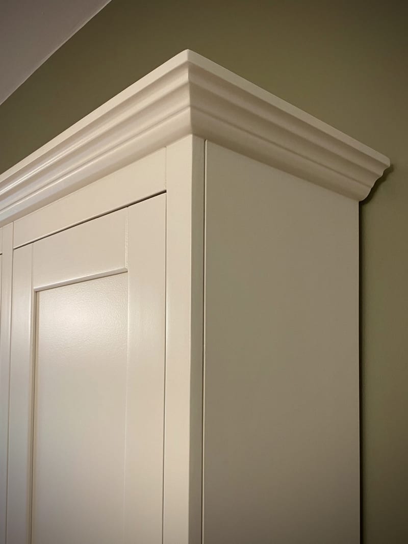 Detail of the top corner, showing the layered moulded cornice and the painted side panel meeting the shaker door against the olive green wall behind