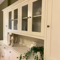 Angled view of the glazed upper cupboards and tall solid end door, with candlesticks, a white two-handled urn and trailing ivy standing on the tongue-and-groove backed shelf below