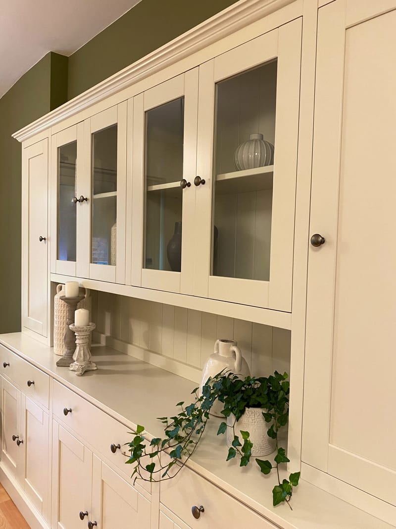 Angled view of the glazed upper cupboards and tall solid end door, with candlesticks, a white two-handled urn and trailing ivy standing on the tongue-and-groove backed shelf below