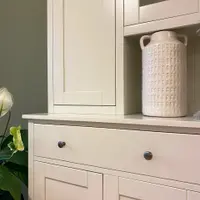 Close view of the left solid upper door and the drawer beneath, showing the shaker panel, aged bronze knobs, a textured cream vase and peace lily leaves in the foreground