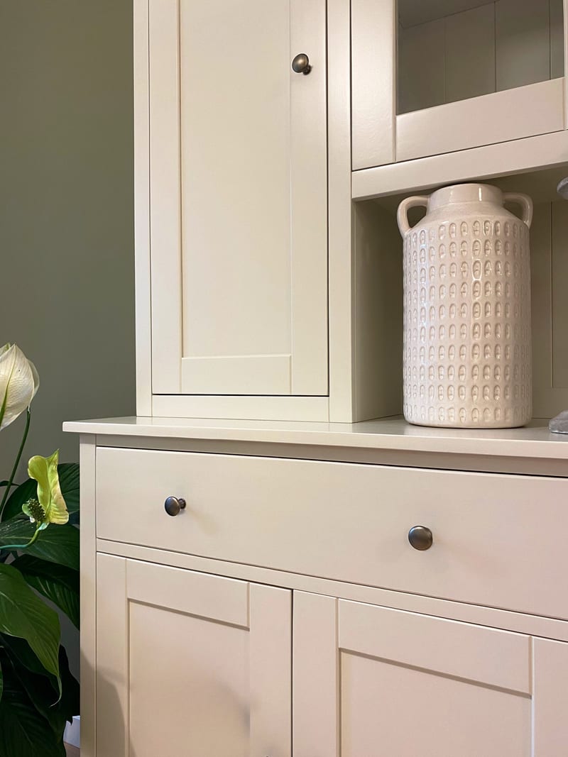 Close view of the left solid upper door and the drawer beneath, showing the shaker panel, aged bronze knobs, a textured cream vase and peace lily leaves in the foreground