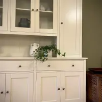 View of the right-hand end, showing glazed doors over shelves of pottery, the tall solid door, two drawers and panelled cupboards, with a carved wooden side table alongside