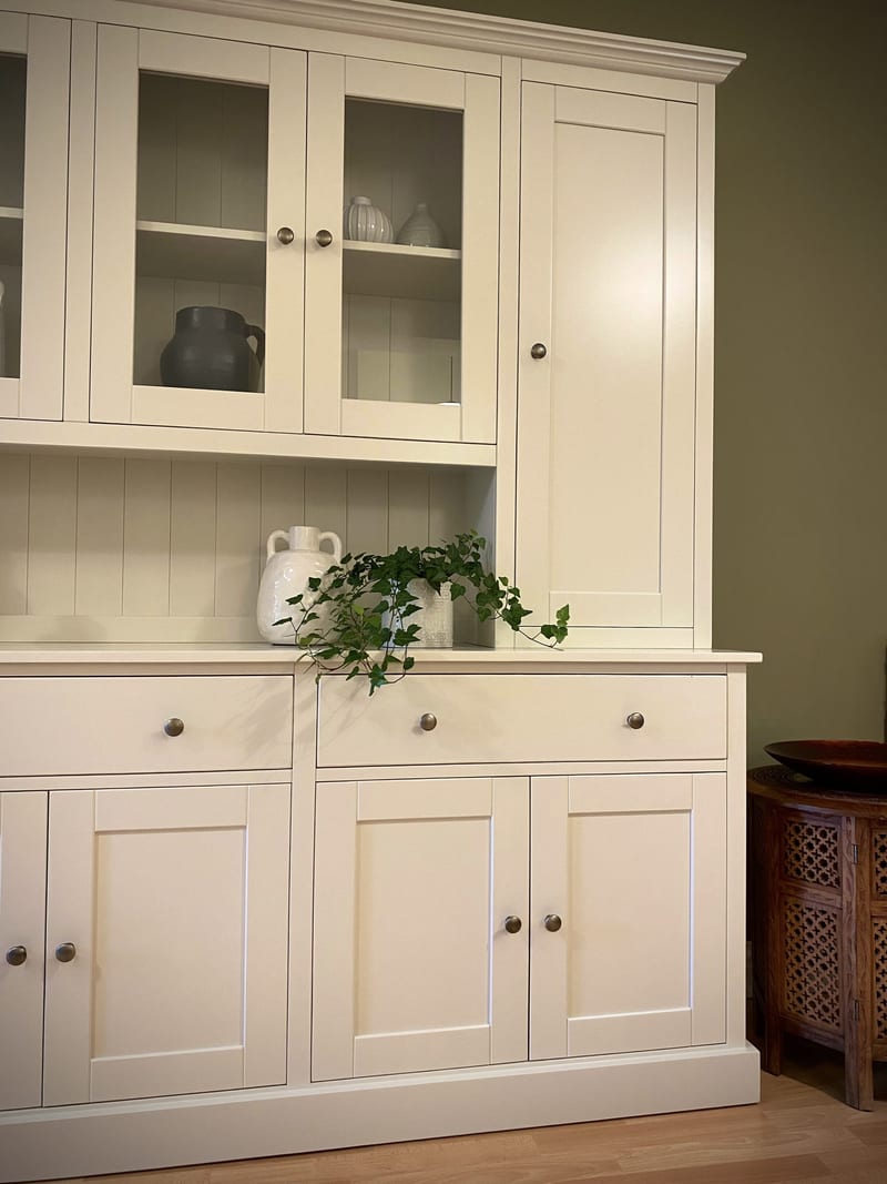 View of the right-hand end, showing glazed doors over shelves of pottery, the tall solid door, two drawers and panelled cupboards, with a carved wooden side table alongside