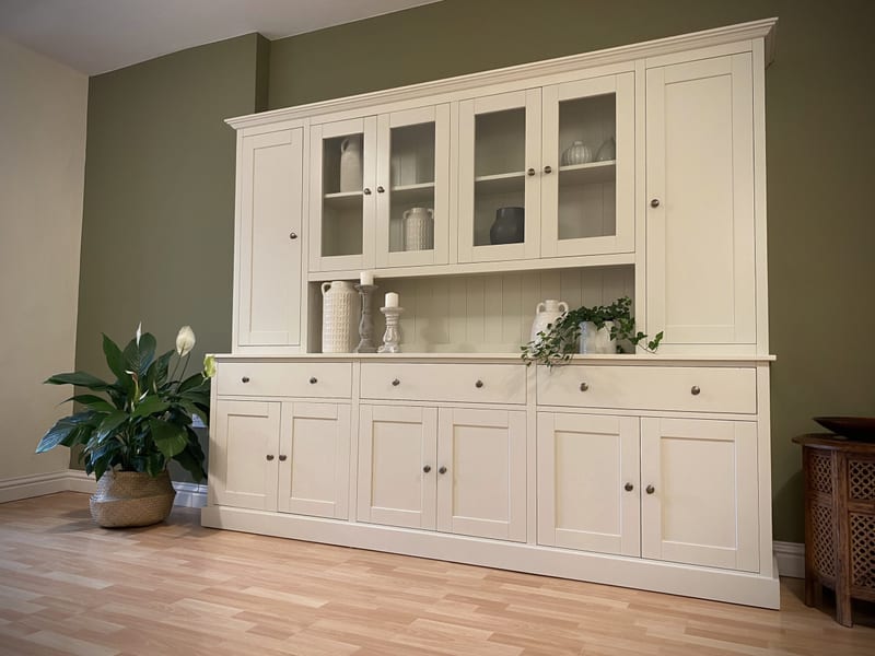 Full front angled view of the cream dresser, showing the glazed and solid upper doors, three drawers, six panelled cupboards and moulded plinth beside a large potted peace lily