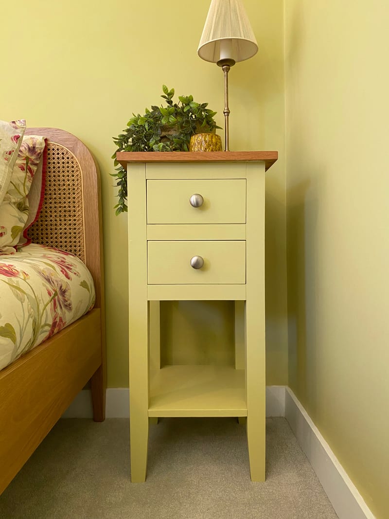 Olive painted small bedside cabinet with a solid oak top, two drawers above an open shelf on tapered legs, beside a rattan headboard with a lamp and a plant on top
