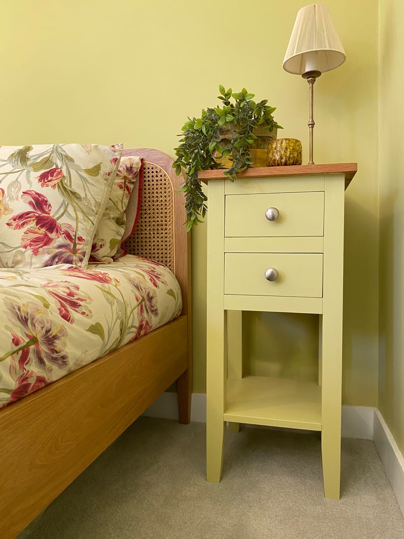 Angled view of the soft yellow painted bedside cabinet with its oak top, two drawers with round brushed nickel knobs and open lower shelf, beside a cane headboard bed with floral bedding