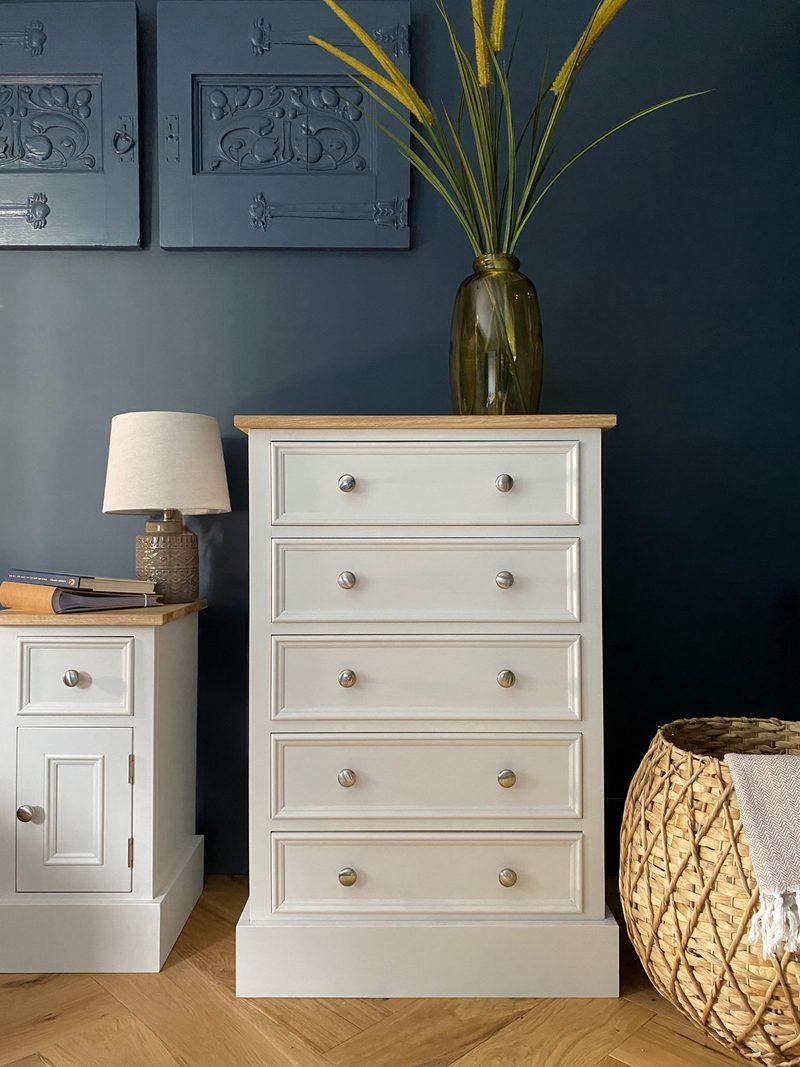 Full front view of the off-white painted five-drawer chest with an oak top and plinth base, brushed nickel knobs and a green glass vase of grasses, set against a dark blue wall