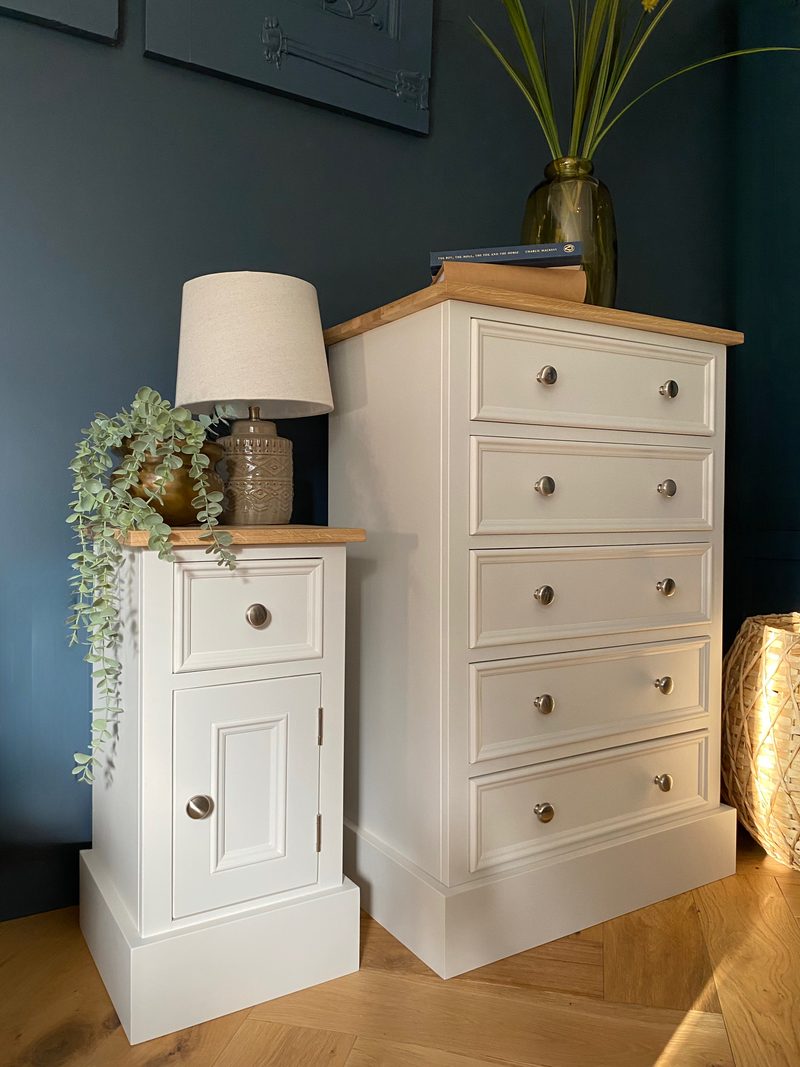 Three-quarter view of the five-drawer chest and a matching small cupboard with a table lamp and trailing eucalyptus, both off-white painted with oak tops on a herringbone oak floor