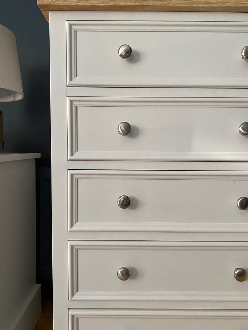 Close view of the upper drawer fronts, showing the deep bevelled mouldings, round brushed nickel knobs and the edge of the oak top, with a lampshade and dark blue wall behind