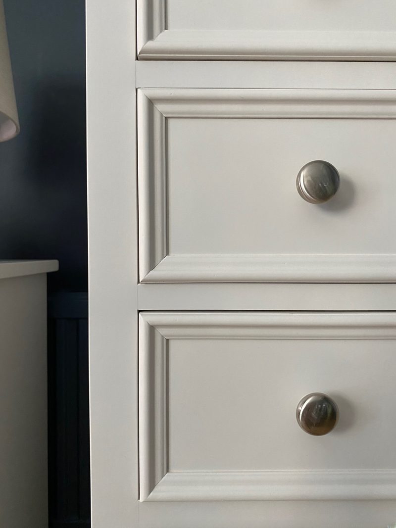 Detail of the front corner of the chest, showing the mitred drawer mouldings, the painted carcase edge and two round brushed nickel knobs against the dark blue wall