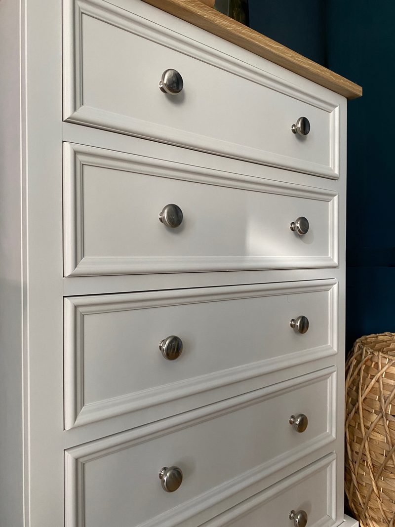 Low angled close view along the drawer fronts, showing the moulded panels, pairs of brushed nickel knobs and the oak top edge, with a woven basket beside the dark blue wall