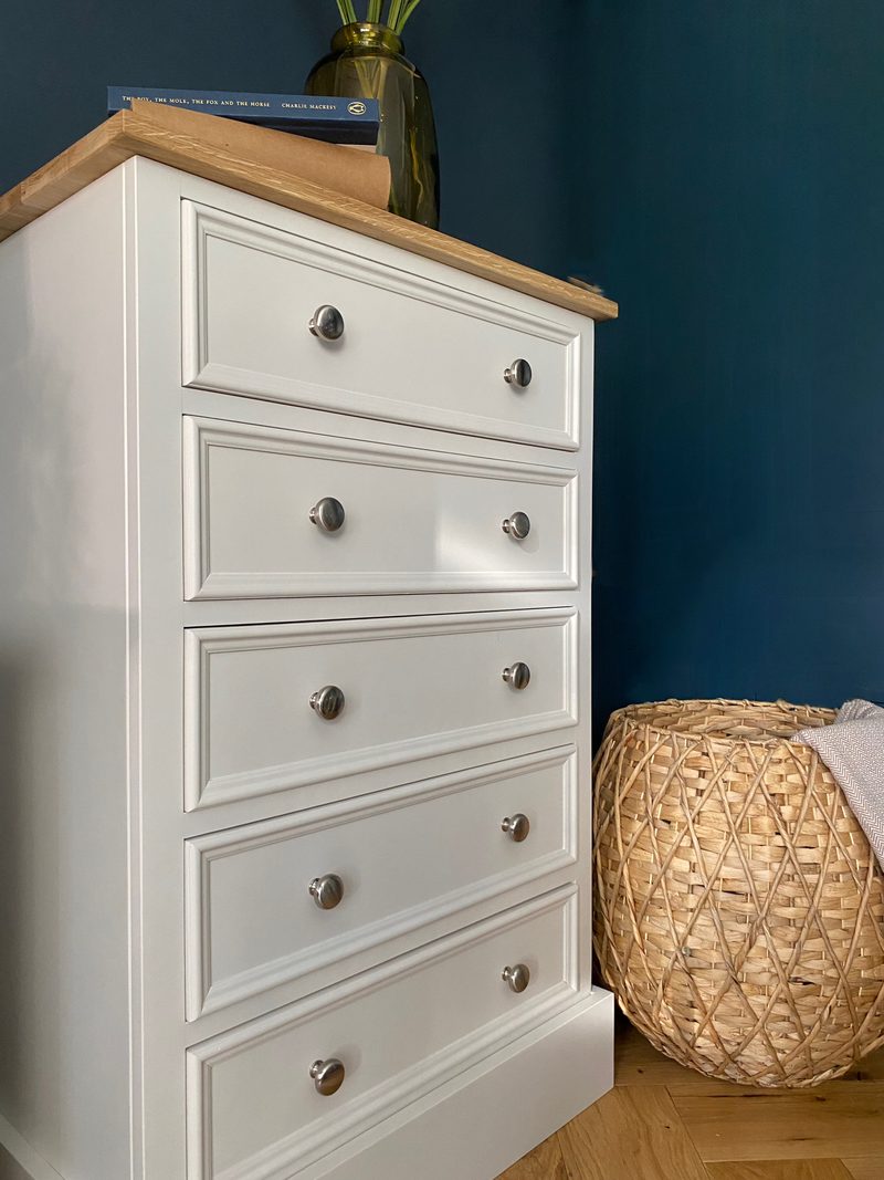 Side angled view of the chest showing the painted end panel, five moulded drawers, oak top with books and a glass vase, and a woven basket of throws on the oak floor