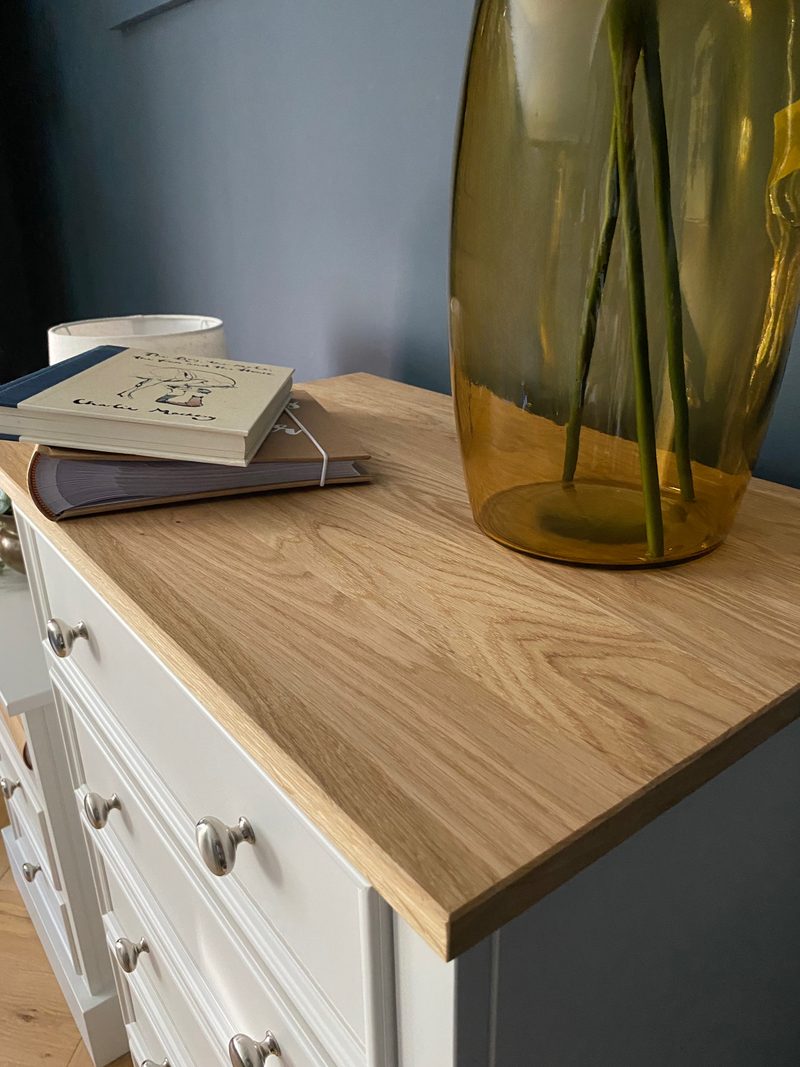 Close view of the corner of the solid oak top, showing the grain and eased edge beside a stack of books and a large olive glass vase, with painted drawer fronts below