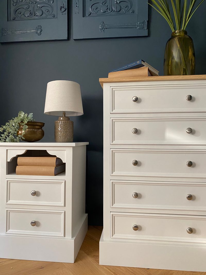 Front view of the five-drawer chest with its oak top beside a smaller painted cabinet holding a lamp, its open recess stacked with books, below carved panels on the dark blue wall