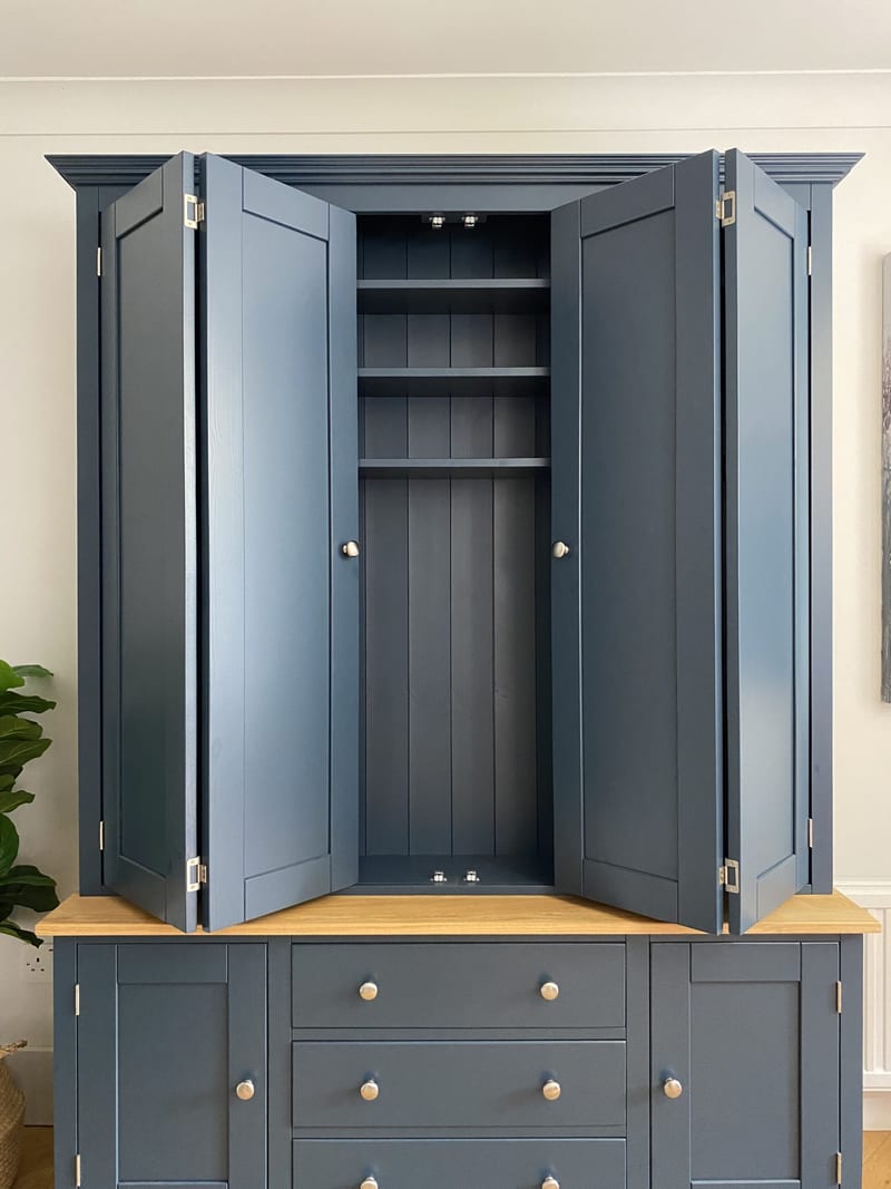 Front view with the bifolding doors folded back, showing the tongue-and-groove lined interior, three adjustable shelves and small spotlights above the oak worktop