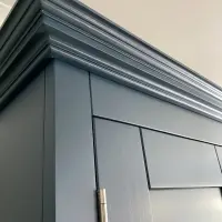 Close detail of the top corner, showing the layered cornice moulding, the crisp navy paint finish and a brushed nickel hinge on the door below, against a cream wall