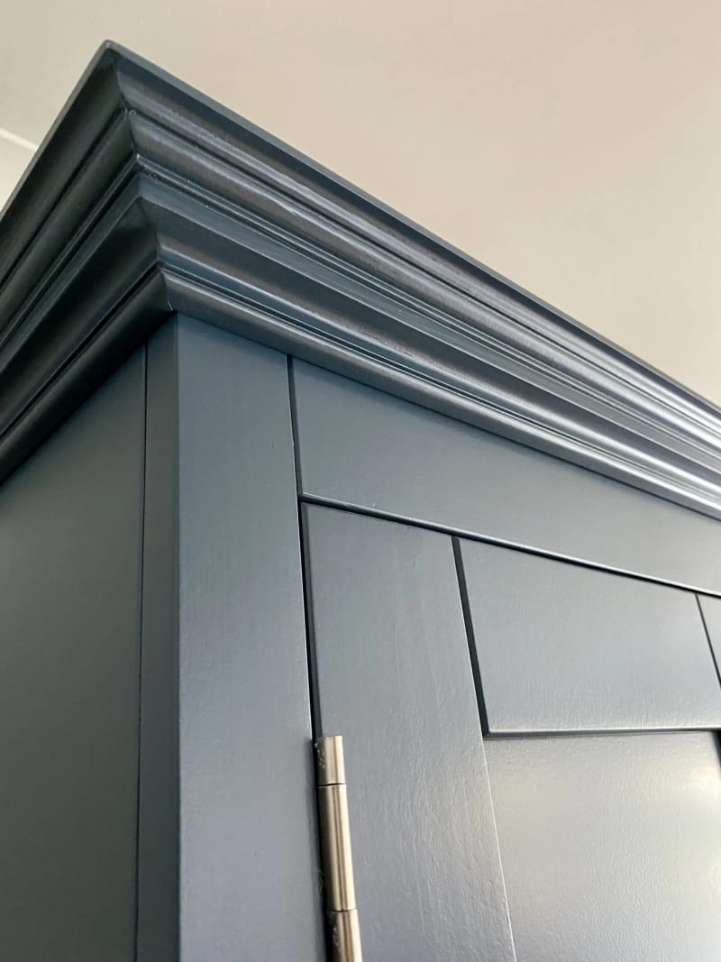 Close detail of the top corner, showing the layered cornice moulding, the crisp navy paint finish and a brushed nickel hinge on the door below, against a cream wall