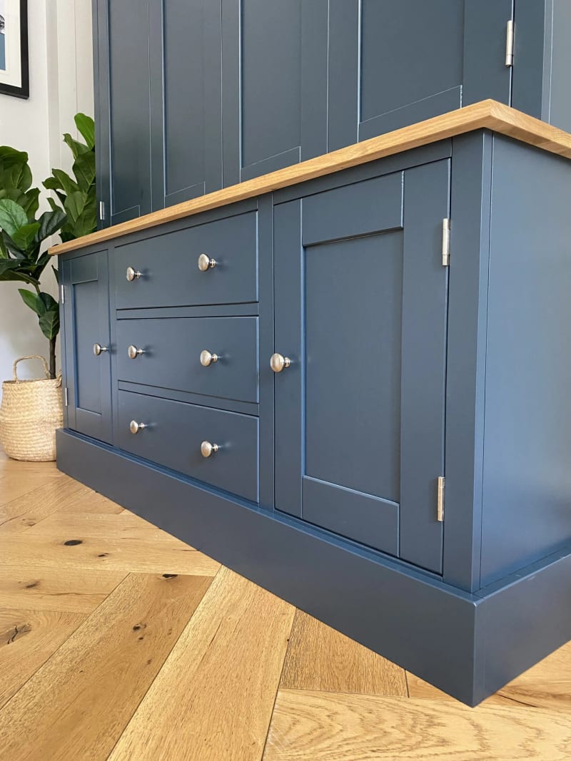 Low angled view of the base, showing three drawers flanked by two shaker cupboard doors, the oak worktop and deep plinth, with a potted plant on the herringbone floor