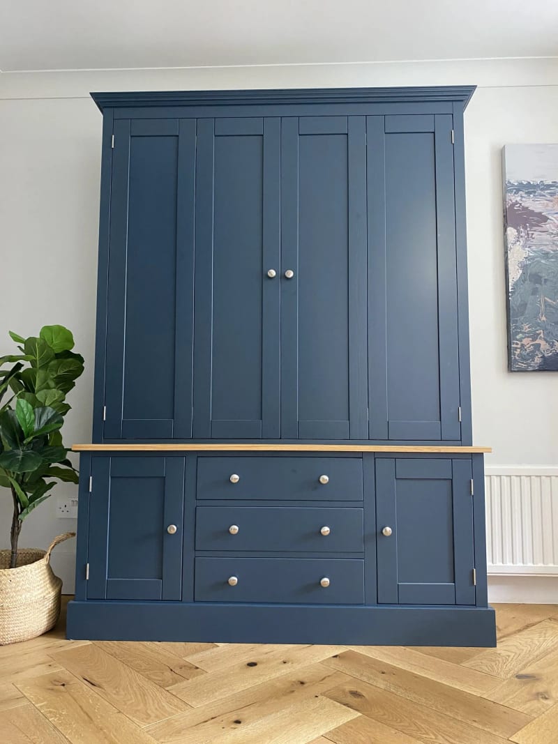 Straight-on view of the navy painted unit with all doors closed, its moulded cornice, oak worktop and brushed nickel knobs, beside a fiddle leaf plant and an abstract painting