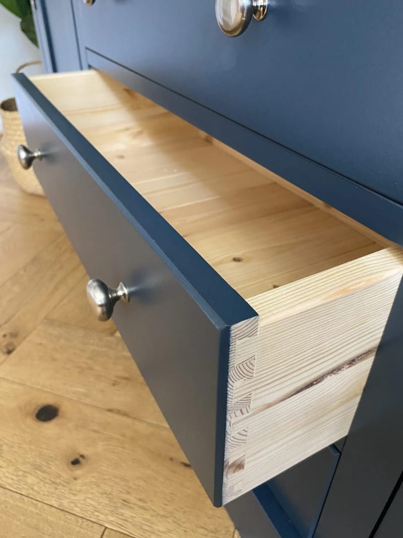 Drawer pulled fully open, showing the natural pine drawer box and hand-cut dovetail joints behind the navy painted front with its round brushed nickel knob, above a herringbone oak floor