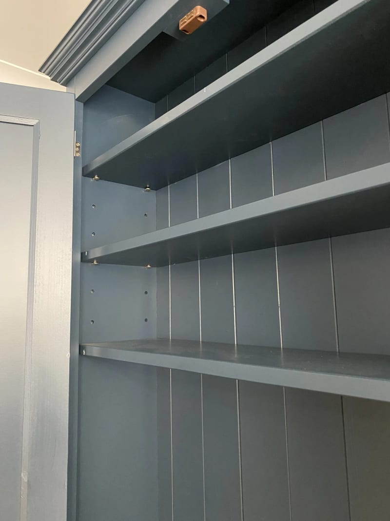 Close view inside the top section, showing three painted shelves on adjustable pegs, the tongue-and-groove back boards and a small copper coloured cable clip at the top